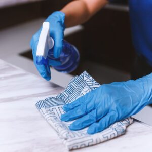 A close-up of gloved hands wiping a kitchen counter with a spray bottle and a blue patterned cloth, representing regular cleaning maintenance.
