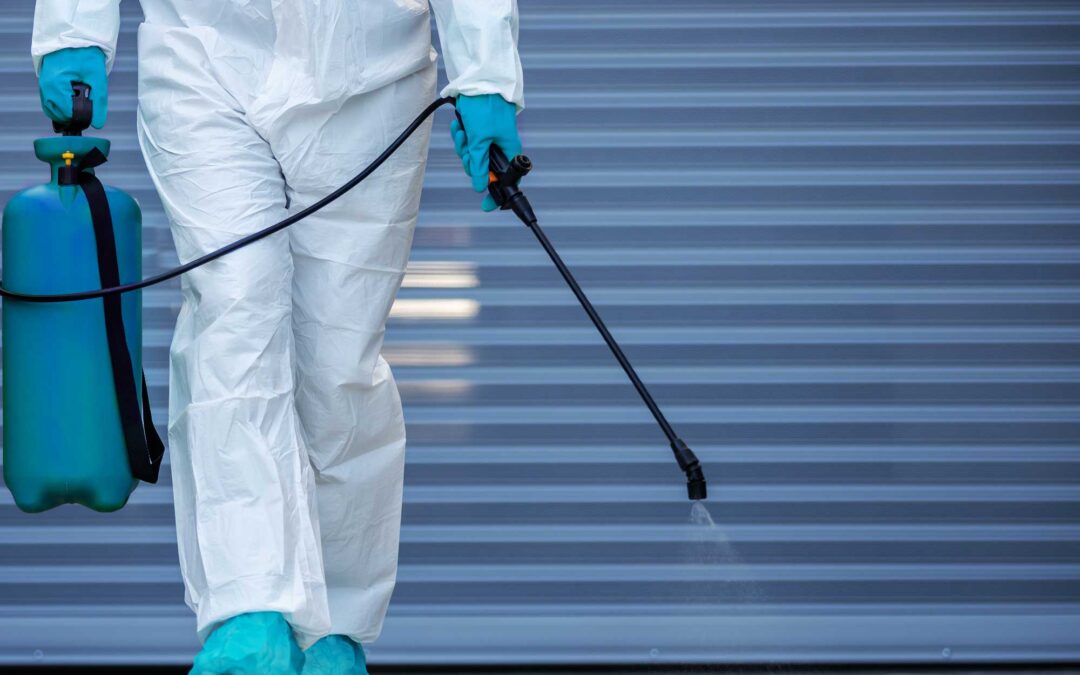 A worker in a full-body protective suit with a respirator is methodically disinfecting the floor using a pressure sprayer, against a backdrop of a metallic corrugated wall, emphasizing cleanliness in sensitive environments.